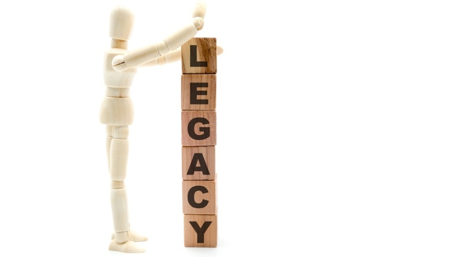Wooden figure as businessman building tower of wooden blocks spelling "Legacy" vertically on white background.