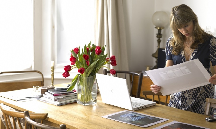 Young woman standing by laptop on dining table looking at printed images.