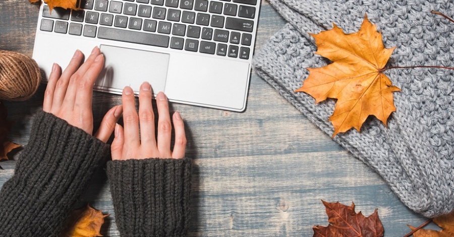 Desk with laptop, fallen leaves on grey wooden background. Flat lay, top view. A woman's hands typing.