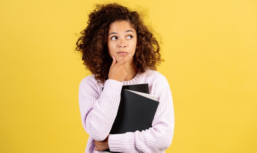 Young woman, looking to the right slightly, thinking with one hand on her chin. She is holding notebooks and folders in her hands, while standing in front of a yellow background.