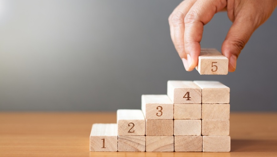 Hand arranging wooden blocks stacking on wooden table in the shape of a staircase. The stack goes up five levels.