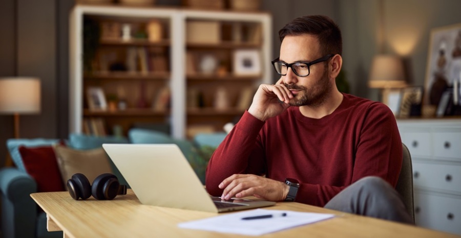 Young male freelancer sitting at a desk and resting his head on his hand while working on a project on his laptop.