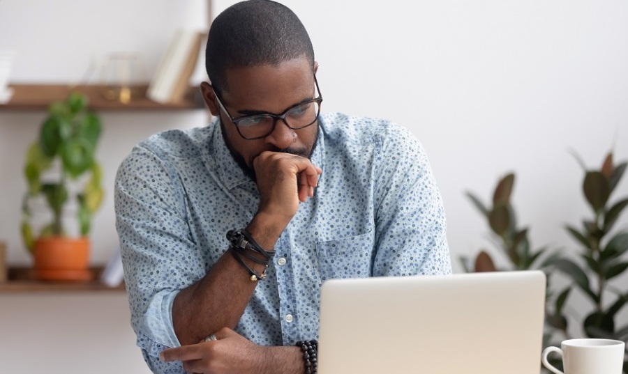 Black guy looking at laptop and thinking with his right hand clasped in front of him.