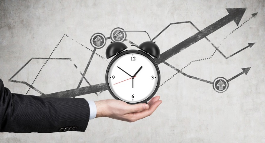 A businessman's hand holding a clock. There is a line chart showing growth behind the alarm clock.