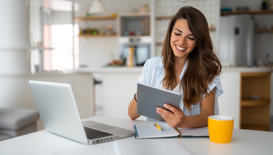 Female business person sitting at table using digital tablet and laptop while working at home.