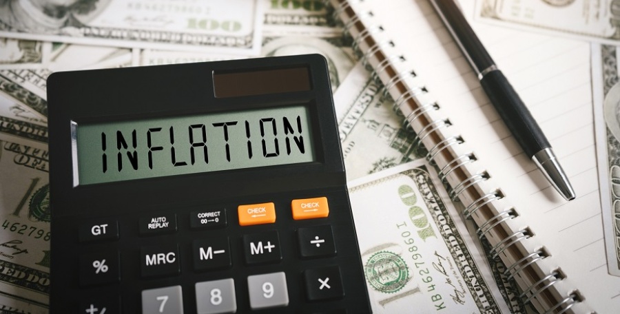 A calculator laying on top of $100 bills with a notebook and pen laying next to it. The word inflation is spelled out on the calculator screen.