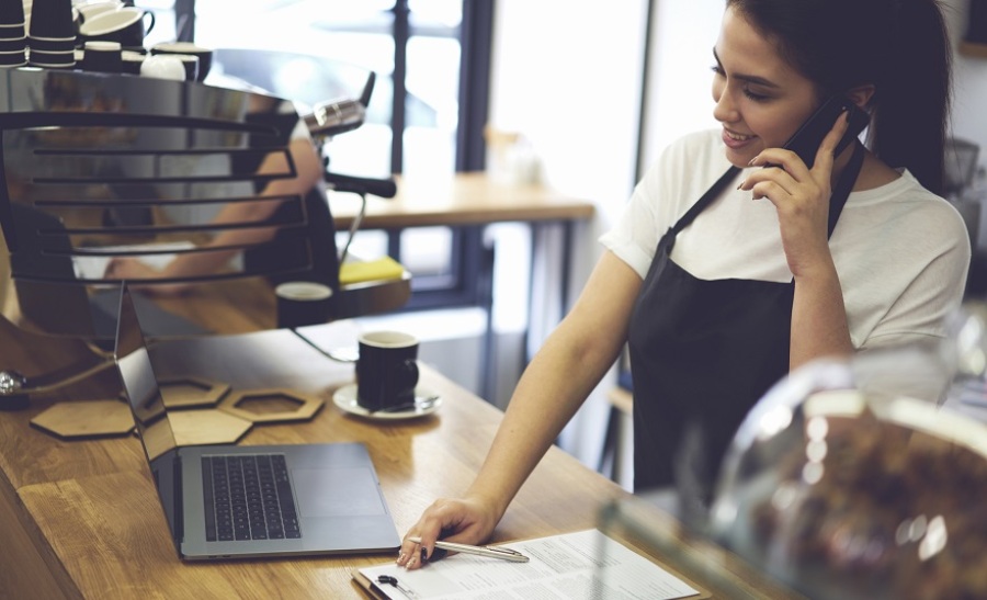 Woman talking on her phone while at the counter of a cafe. A laptop and business report are on the counter.