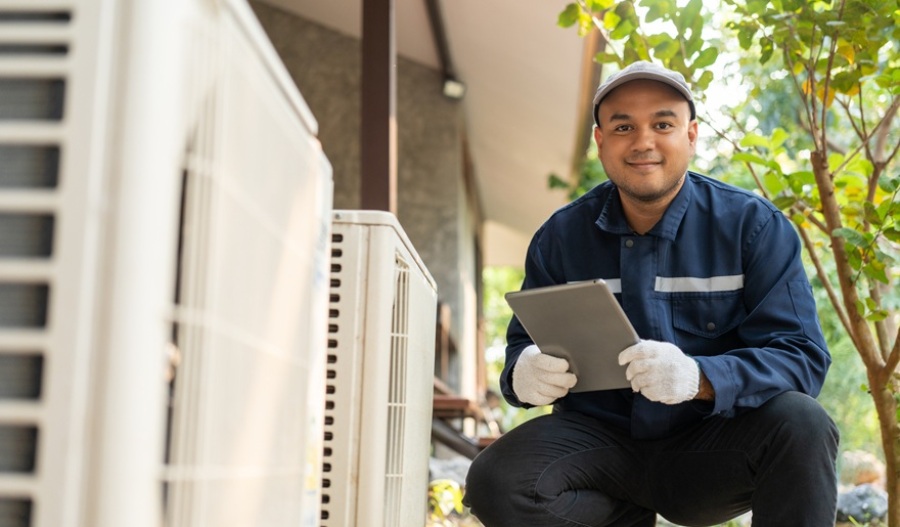 HVAC technician checking outdoor unit. He has a clipboard in his hands and is grinning at the camera.