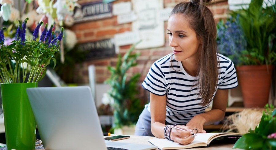 Young female florist writing in notebook and looking at laptop while leaning on counter.