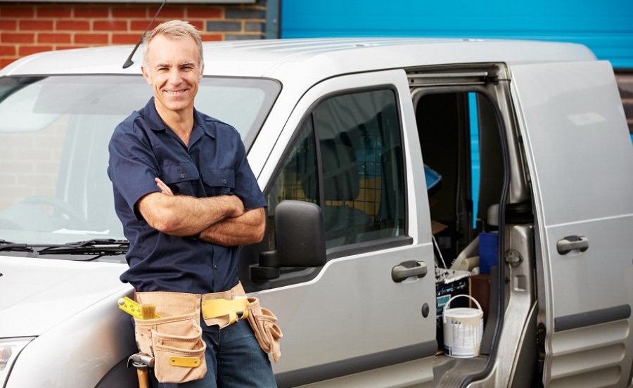 Man with tool belt around his waist leaning against the hood of his gray service van.