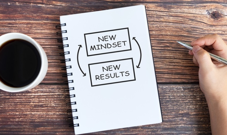Journal and cup of coffee sitting on wooden table. Entry in journal reads "New Mindset, New Results". A person's hand holding a pen is on the right of the image.