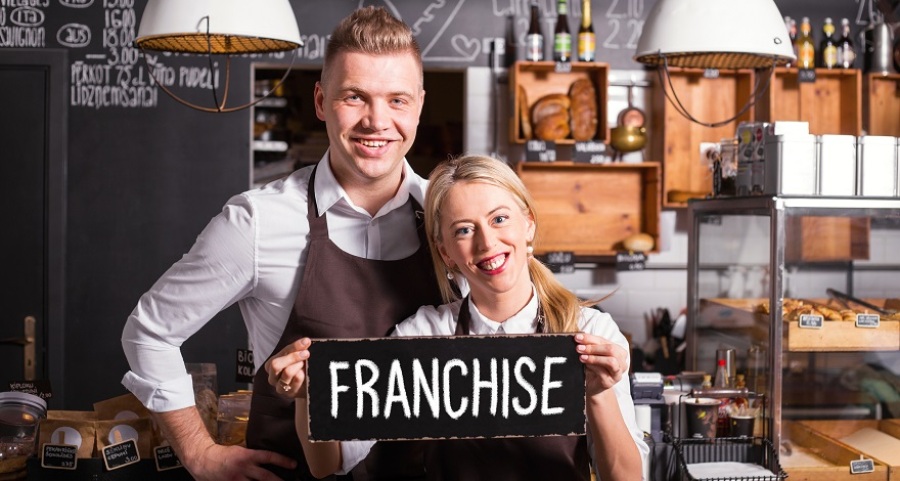 Man and woman in a bakery or café smiling at the camera while the woman holds up a sign saying "Franchise".