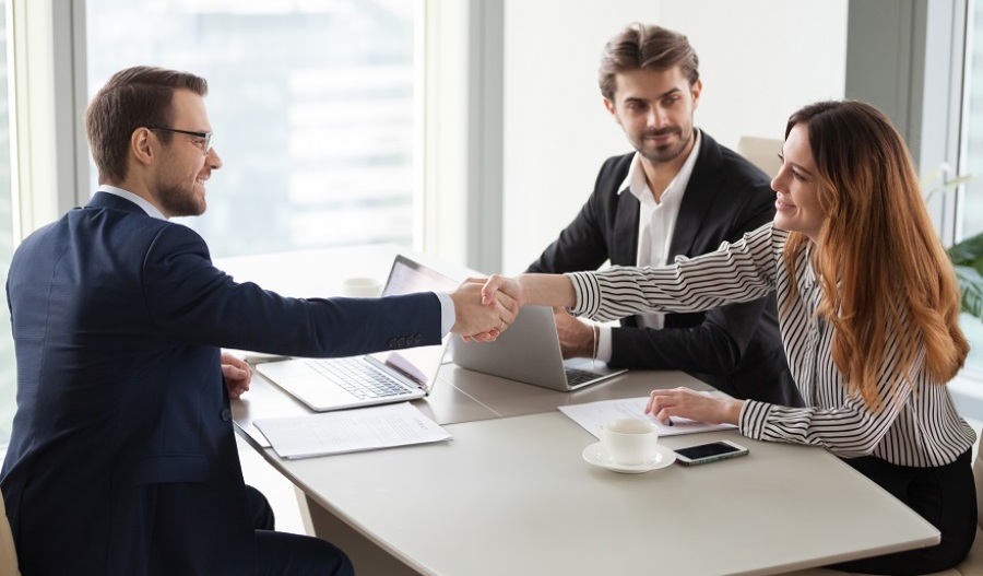Businessman and businesswoman shaking hands over desk after finishing group negotiations. Additional businessman sits smiling in background next to the businesswoman.