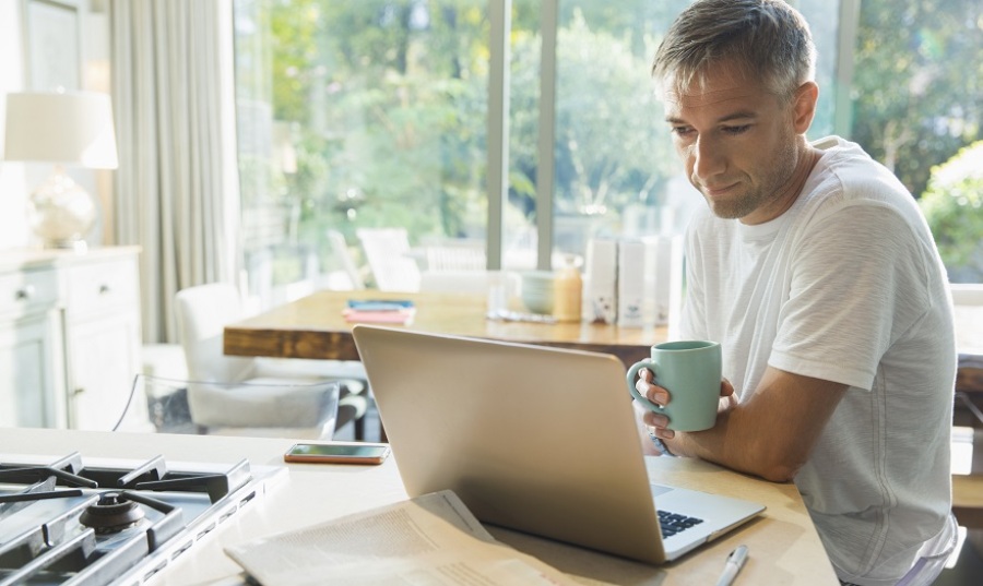 Man sitting at a kitchen counter drinking coffee and working on a laptop.