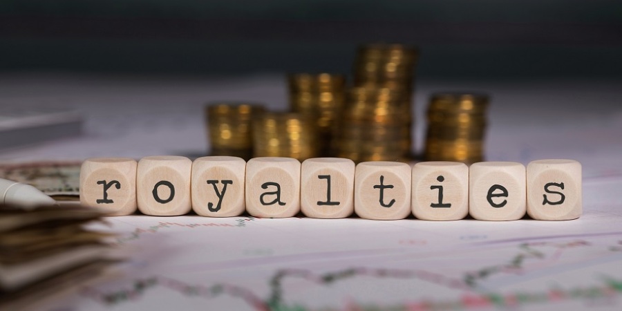 The word royalties spelled out with wooden blocks on top of papers with financial graphs and stacks of coins in the background.