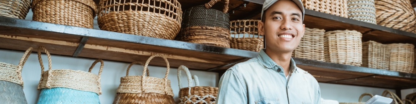 Photo of smiling young male entrepreneur looking at the camera while holding a digital tablet while standing with a wall of hand-crafted bags in the background.