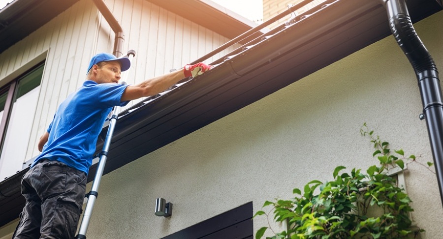 Man in a blue shirt on ladder cleaning house gutters of leaves and dirt.