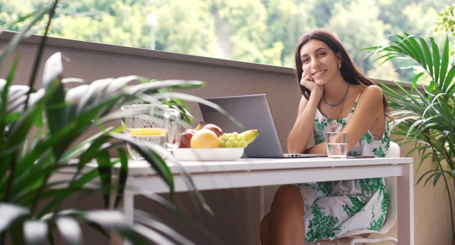 Young woman sitting at a table on a balcony. She is looking at the camera and smiling. There is a laptop open in front of her on the table.