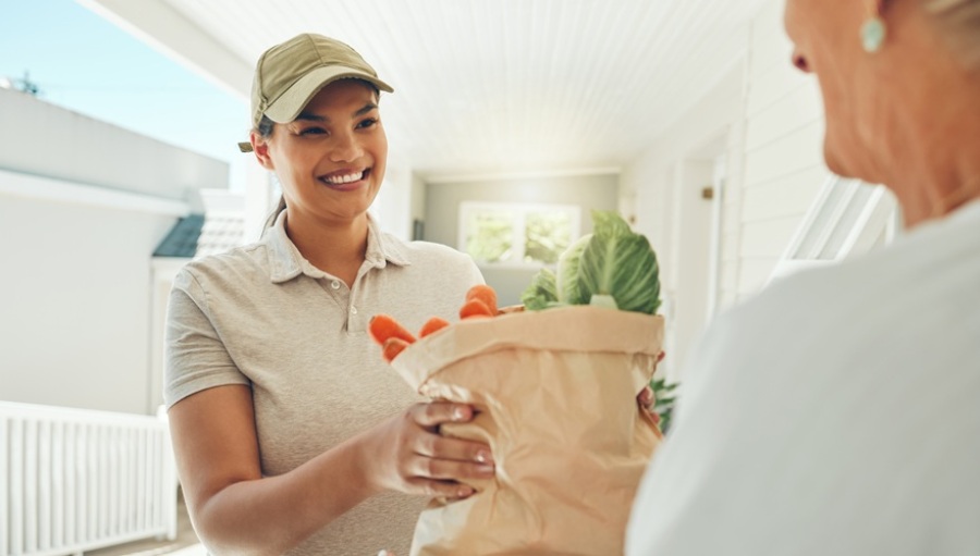 Younger woman smiling and delivering a paper grocery bag to an older woman.