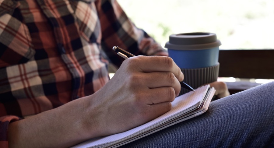 Closeup of a young man wearing jeans and a plaid shirt writing with a pen in a notebook in the porch of a house or a cafe.