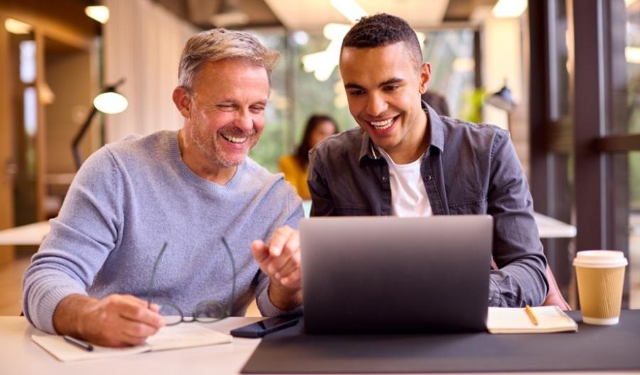 Businessman mentoring younger colleague. Both are seated at a desk looking at a laptop.