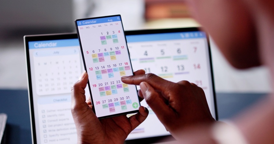 Black woman looking up calendar on cellphone. A shot of calendar is also on the laptop screen in the background.